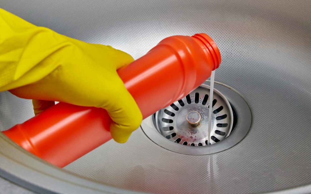 Person's hand in a yellow rubber glove pours pipe cleaner down the drain of a metal kitchen sink. Prevention and cleaning of sewer blockages